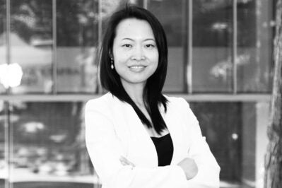 Black and white headshot of a person wearing a light-colored blazer, standing with arms crossed and smiling, in front of a glass wall.