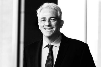 Headshot of a person wearing a suit and tie, smiling. The image is in black and white, with a light background and contrasting dark areas.