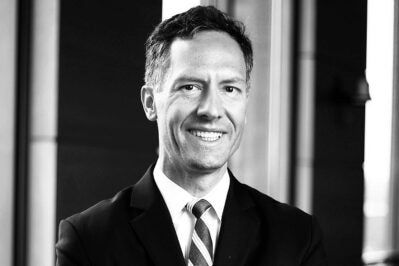 A black and white headshot of a smiling person in a suit and tie, standing in front of a blurred background.