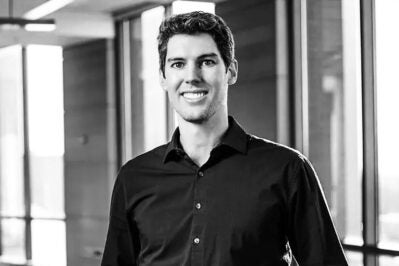 A person smiling in a professional headshot, wearing a dark shirt, with a background of large windows and a softly lit interior.