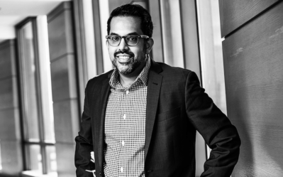 A black and white headshot of a person in a suit and checkered shirt, leaning against a wall in a modern office setting.