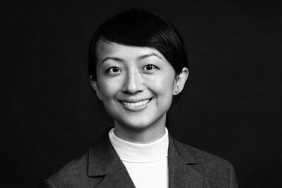 Black and white headshot of a person smiling, wearing a suit jacket and a white turtleneck, set against a dark background.