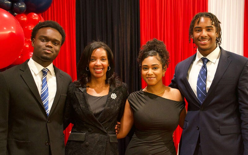 A small group of people posing together, dressed formally in suits and dresses, standing in front of colorful balloons and curtains.
